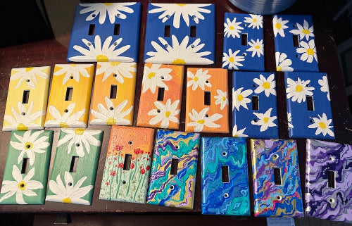 painted light switch plates with white daisy designs on blue backgrounds, yellow backgrounds, orange backgrounds, and green backgrounds, plus covers with multicolored metallic designs, painted by ©Lisa Johnson Arts, Norwich Vermont USA